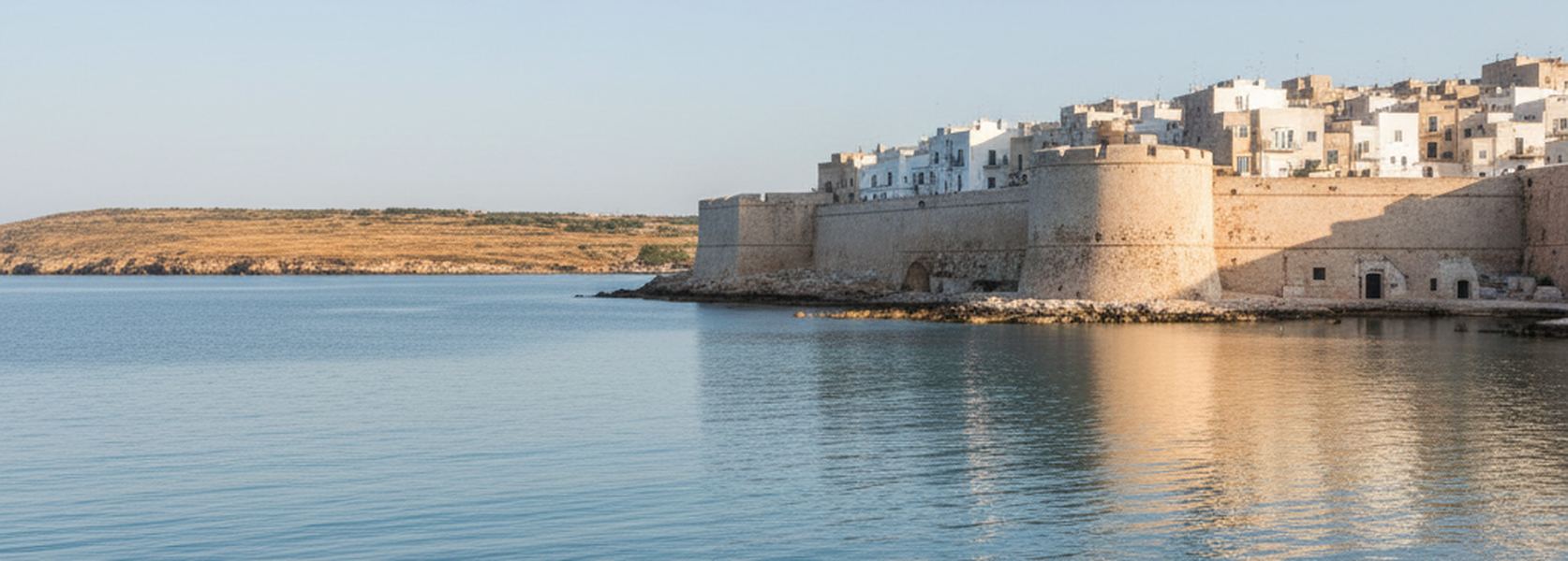 The walled seafront of Otranto seen from the water, pale limestone bastions rising off a shallow rock shelf with the old town stacked behind them.