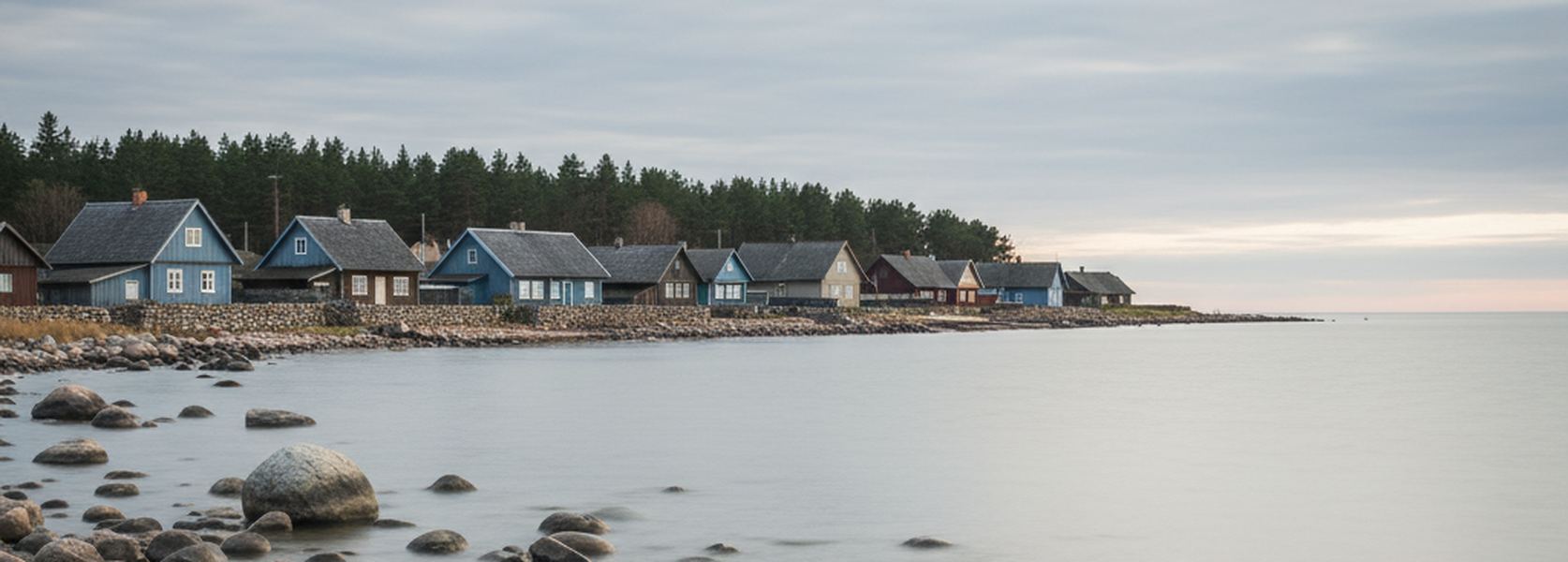 Wooden houses behind low stone walls above a shore of gray boulders in a small Estonian coastal village under flat cloud.
