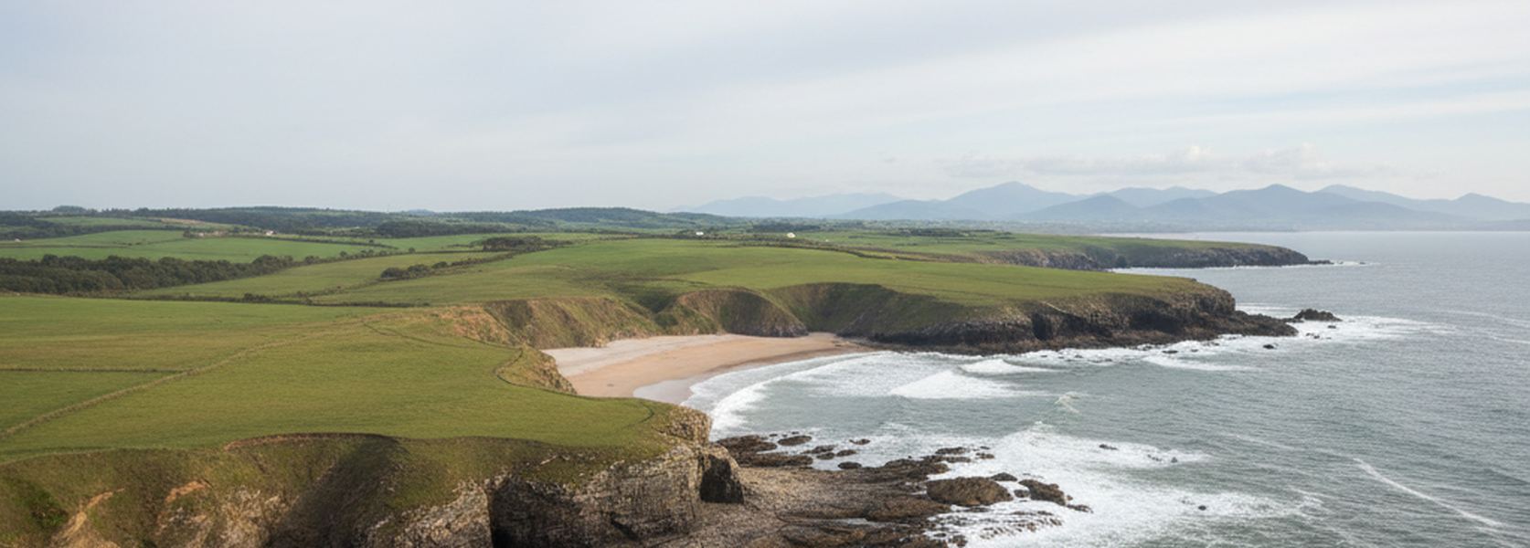 Green pasture ending at low limestone sea cliffs on the north coast of Spain, with a narrow sand cove between two headlands and swell breaking on rock shelves.