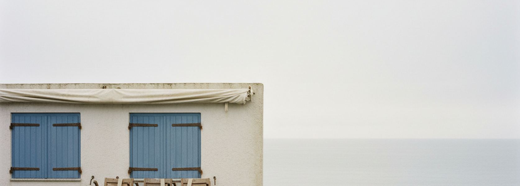 A rendered guesthouse wall with two closed blue shutters and a slack canvas awning, flat sea and pale sky beyond.