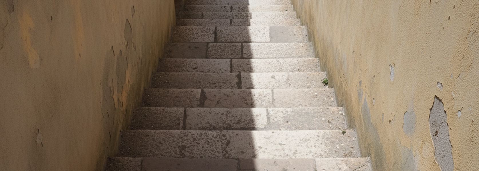 A narrow stepped alley between rendered walls, worn treads with a hard diagonal shadow line running across them.