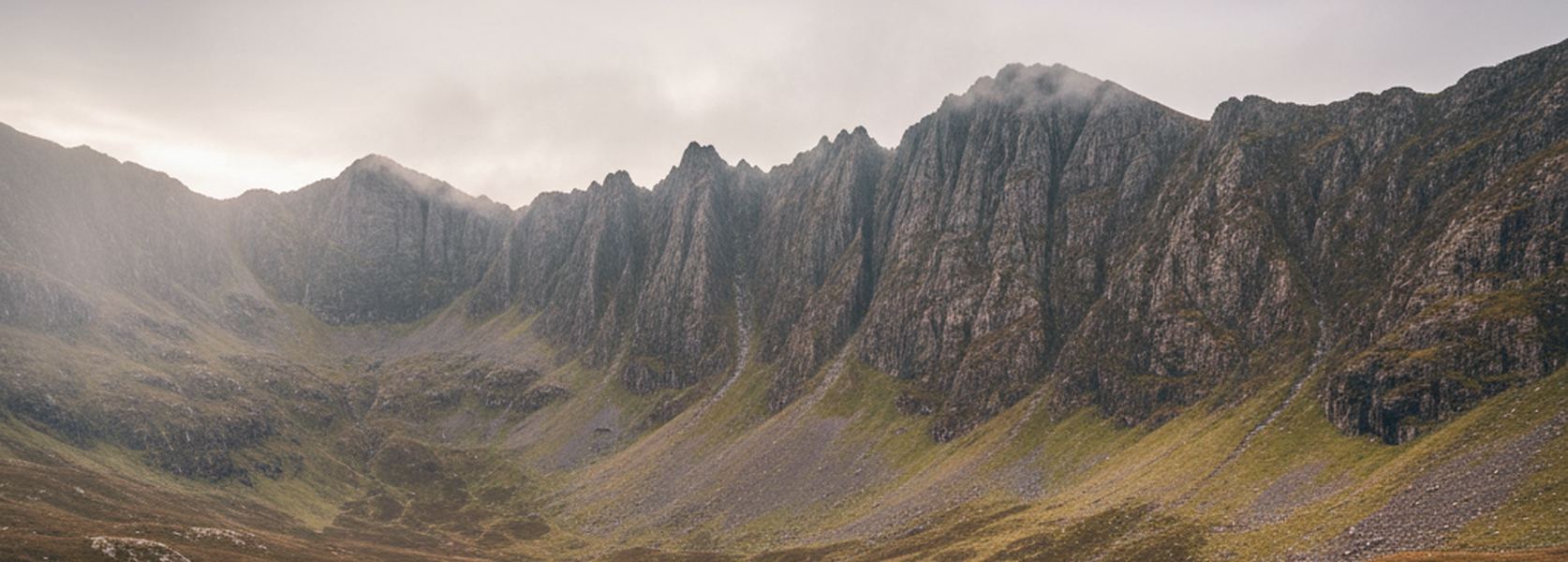 A wide glacial cirque of jagged rock pinnacles above steep grass and scree, with cloud catching the highest summit.