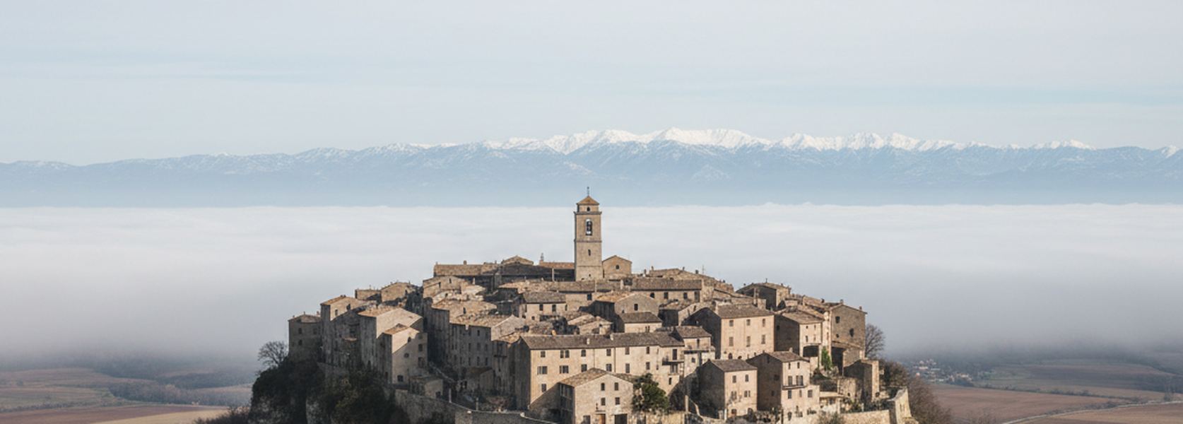 A stone hill town on a rock spur above a sea of valley fog, snow covered mountains along the far horizon.
