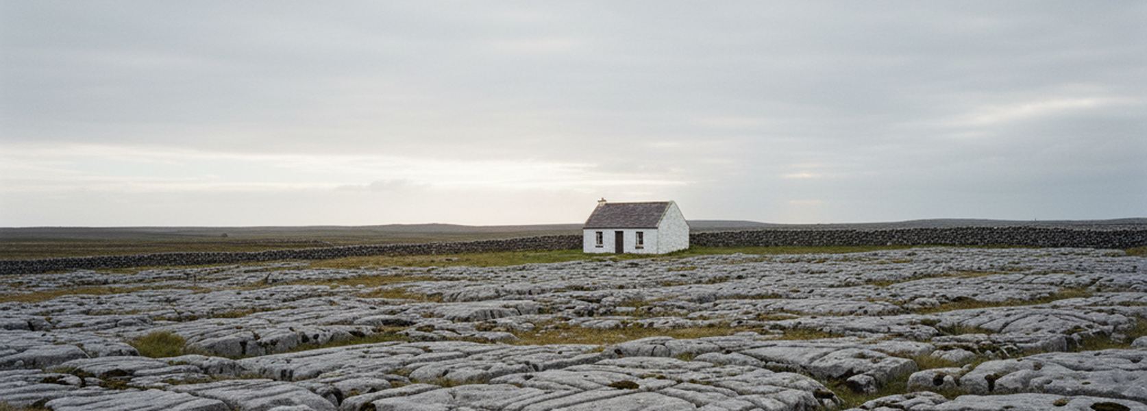 A single small stone cottage standing alone on stepped limestone pavement, one field wall running past it, no road and no other building in sight.