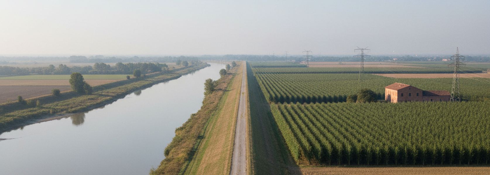 The mown gravel crown of a flood levee running straight between the river and flat fields on the Po plain in September.