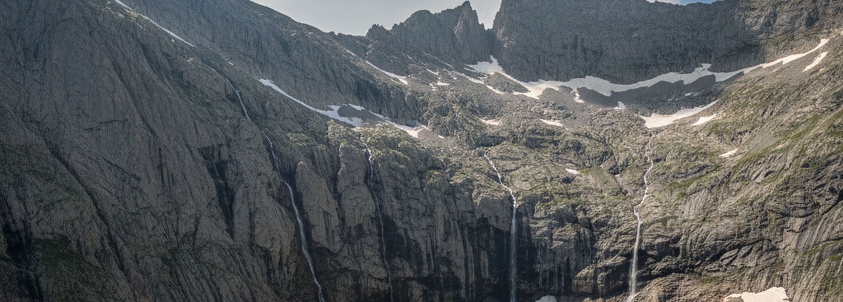 A gray cirque headwall with several thin waterfalls dropping past broad banks of old snow lying on stony terraces.