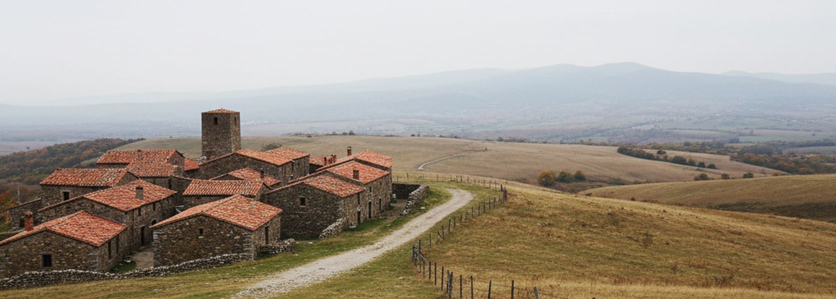 A small settlement of tiled roofs and a squat tower on the left, one lane leaving it, and open pasture behind a wire fence filling the rest of the frame.